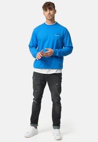 Sudadera azul con cuello redondo y logo bordado. Combinada con jeans negros desgastados y zapatillas blancas, que cuentan con un dobladillo ajustado.