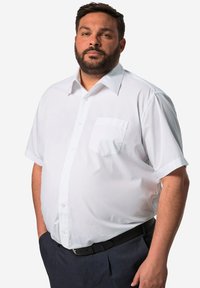 Camicia bianca a maniche corte realizzata in tessuto morbido, con chiusura a bottoni e una tasca singola sul lato sinistro. Cintura nera e pantaloni scuri.