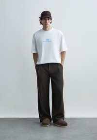 Balti trumpomis rankovėmis marškinėliai su mėlynu grafinio teksto raštu, derinami su laisvai krentančiomis tamsiai rudos spalvos kelnėmis ir rudais raštuotais batais.