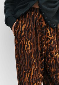 Hnedo-čierne nohavice so zvieracím vzorom a jemnou textúrou, s širokým elastickým pásom a bočnými vreckami; nosené s tmavým kardigánom.