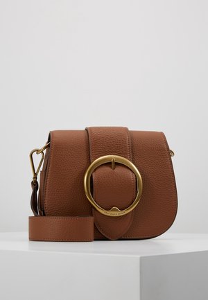 Sac bandoulière en cuir marron texturé avec une large sangle et une grande boucle ronde dorée sur le rabat avant, posé sur une surface blanche.
