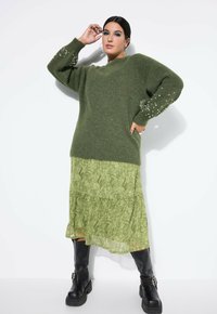 Maglione verde oversize con dettagli di perle sulle maniche, abbinato a una gonna a strati in stile paisley verde e stivali neri fino al ginocchio.
