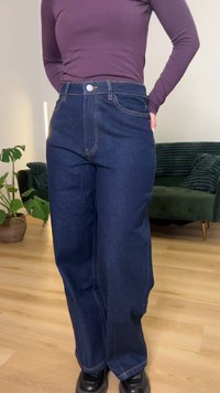 Pessoa vestindo jeans de cintura alta e pernas largas azul escuro com uma blusa de mangas compridas roxa, de pé em piso de madeira perto de um sofá verde e uma planta.