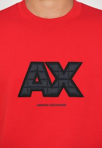 Červené bavlnené tričko s veľkým čiernym prešívaným logom "AX" a textom "ARMANI EXCHANGE" pod ním v čiernej farbe. Okrúhly výstrih a krátke rukávy.