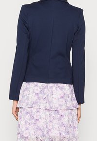 Žena v přiléhavém námořnickém blejzru, který má na sobě volnou světle fialovou květinovou šaty s různými vrstvami, zachycena zezadu na jednobarevném pozadí.