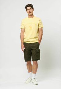 Tricou galben din bumbac cu logo-ul U.S. Polo Assn., asortat cu pantaloni scurți cargo verzi închiși, care au buzunare laterale, purtați cu adidași și șosete albe.