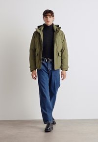 Oliven grøn polstret jakke med lynlås, to sidelommer, båret over en sort turtleneck, parret med løse blå denim jeans og sorte støvler.
