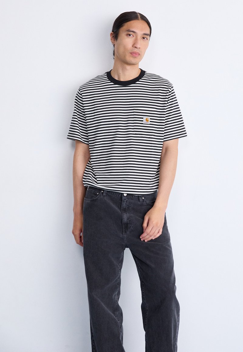 Mand med langt, mørkt hår iført en sort-hvid stribet t-shirt med korte ærmer og løse, mørkegrå jeans, stående mod en ensfarvet hvid baggrund.