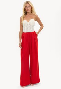 Femme aux longs cheveux blonds portant un corset blanc en dentelle sans bretelles et un pantalon rouge taille haute à jambes larges, debout avec une main dans la poche.