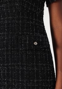 Svart tweedklänning med en strukturerad yta, prydd med silverdetaljer och en framficka med dekorativ knappdekoration.