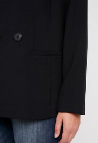 Primer plano de una persona vistiendo un blazer negro con detalle de botón y bolsillo, combinado con jeans de mezclilla azules, con la mano relajada al costado.