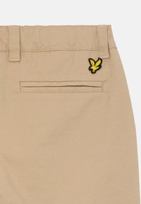 Beige bukser med en baglomme med sort og gul ørner broderi. Materialet ser glat ud med en subtil tekstur.