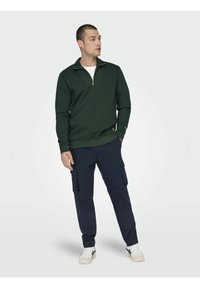 Felpa verde scuro con zip a quarto e polsini a coste, abbinata a pantaloni cargo blu navy con multiple tasche e scarpe da ginnastica bianche.