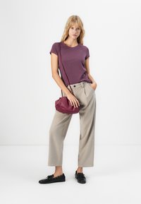 Blonde Frau, die ein lila T-Shirt, beigefarbene weite Hose, schwarze Slipper trägt und eine kleine lila Umhängetasche hält, vor einem weißen Hintergrund steht.