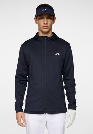 Marineblå zip-jakke med hætte, der har et hvidt logo på brystet, struktureret stof og slim fit-design. Bliver konsekvent parret med hvide bukser.