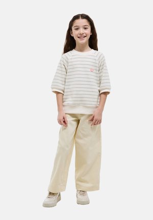 Fille souriante aux longs cheveux foncés portant un sweat-shirt blanc à rayures, un pantalon large crème et des baskets beige clair, debout sur un fond blanc.