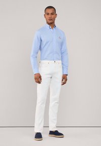 Lichtblauwe button-up shirt met lange mouwen, witte denim broek en marineblauwe espadrilles, met een klein geborduurd logo op het shirt.