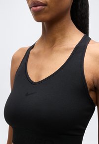 Femme portant un soutien-gorge de sport Nike noir avec un col en V, montrant le cou, les épaules et le haut de la poitrine sur un fond uni.