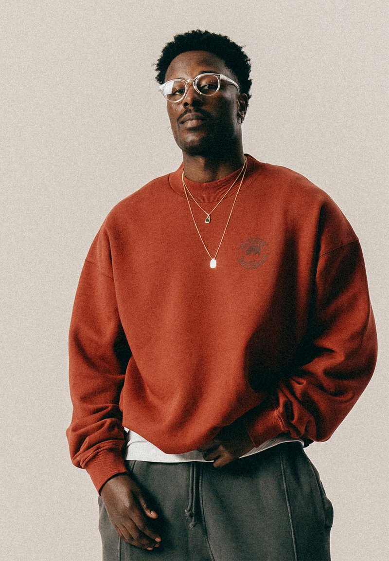 Άνδρας με κοντά σγουρά μαλλιά και γυαλιά, φορώντας δύο χρυσά κολιέ, κόκκινο φούτερ και γκρι παντελόνι, στέκεται μπροστά από μία απλή κλασική φόντο.