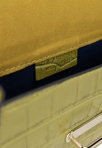 Zelená taška vyrobená z pravé kůže s krokodýlím vzorem, zlatými komponenty a žlutým semišovým interiérem s modrým podšitím.