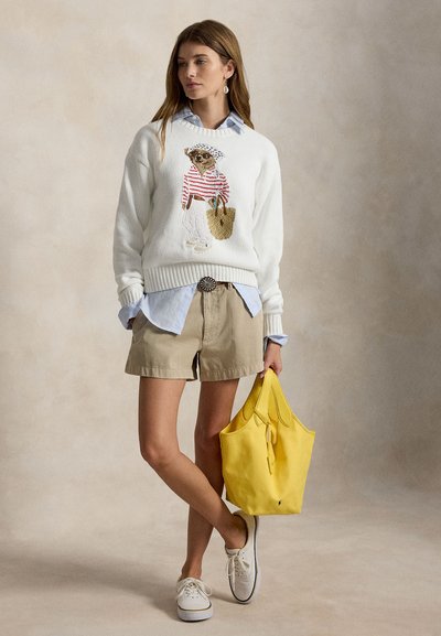 Γυναίκα που φοράει λευκό πουλόβερ με εικόνα αρκούδας, μπλε πουκάμισο, χακί σορτς, λευκά αθλητικά παπούτσια, κρατώντας μια μεγάλη κίτρινη τσάντα ώμου, στέκεται μπροστά σε μπεζ φόντο.