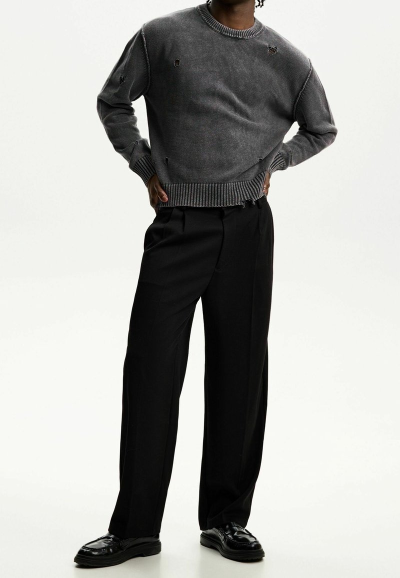 Szary dziany sweter z przetarciami i prążkowanymi mankietami, w połączeniu z czarnymi szerokimi spodniami i błyszczącymi czarnymi butami.