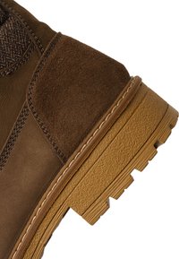 Detailní záběr paty hnědé kožené a semišové boty s propracovaným stehováním a texturovanou pryžovou podrážkou.