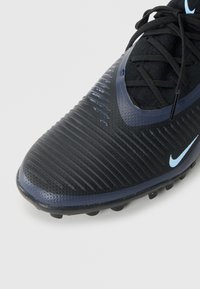 Botas de fútbol Nike con un upper negro texturizado, acentos azules y un diseño aerodinámico. Presenta una puntera redonda y relieves elevados para un mejor agarre.