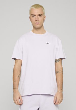 Ung mann med kort blondt hår iført en enkel hvit oversized t-skjorte med "UNFAIR"-logo på brystet og synlige tatoveringer på armene.