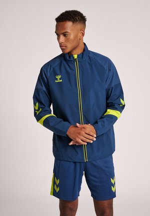 Marine sportsjakke med gule accenter; frontlynlås, høj krave og logo på brystet. Sammen med marine shorts med et chevronmønster.