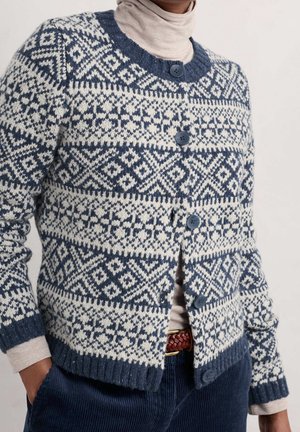 Osoba w niebiesko-białym swetrze w wzory na guzikach, nosząca beżowy golf i niebieskie spodnie z brązowym plecionym paskiem.