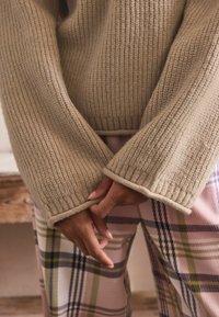 Beige kötött pulóver széles bordás ujjakkal, rózsaszín kockás nadrággal párosítva, amelyen átlós zöld és lila csíkok találhatók. Textúrázott és rétegezett dizájn.