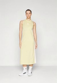 Kvinne som står og har på seg en lys gul ermepotet midi-kjole med hvite sidestriper, hvite sokker og hvite Adidas joggesko.