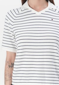 Hvit kortermet polo-skjorte med marineblå horisontale striper, med V-hals og liten logo på brystet. Myk, glatt tekstilfinish.