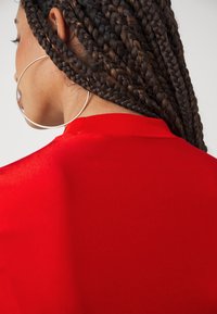 Κόκκινο κοντομάνικο πουκάμισο με στρογγυλή λαιμόκοψη, που διαθέτει μια λεία υφή. Μεγάλο χρυσό σκουλαρίκι σε σχήμα κρίκου ορατό στα πλεξίματα των μαλλιών του μοντέλου.