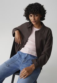 Donna con capelli ricci che indossa una giacca con cerniera marrone scuro, top rosa chiaro e jeans blu, seduta con un braccio appoggiato su una sedia.