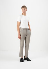 Ung mand står, iført en hvid T-shirt, beige skræddersyede bukser og sorte læder loafers, og kigger til siden mod en ensfarvet baggrund.