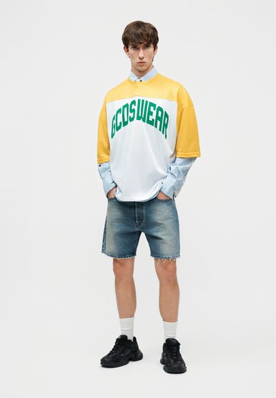 Νεαρός άνδρας που φοράει κίτρινη και λευκή μπλούζα GCDSWEAR πάνω από μπλε ριγέ πουκάμισο, τζιν σορτς, λευκές κάλτσες και μαύρα αθλητικά παπούτσια, όρθιος με τα χέρια στις τσέπες.