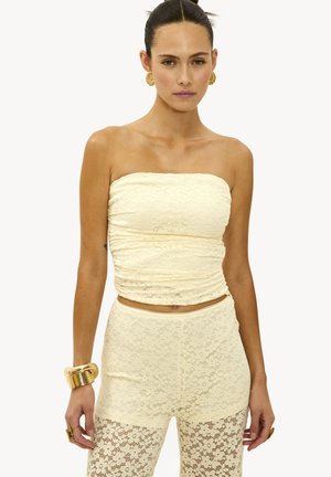 Femme portant un haut en dentelle sans bretelles beige et un pantalon en dentelle assorti, accessorisé avec des boucles d'oreilles dorées audacieuses, un bracelet et des bagues sur fond blanc.