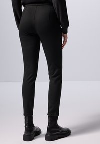 Černé slim fit kalhoty s hladkým materiálem, navržené s vysokým pasem a zúženými nohavicemi. Nosené s černými kotníkovými botami.