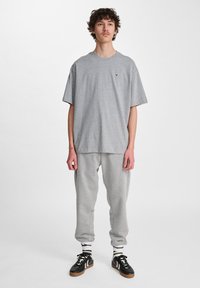 Camiseta de algodón gris con cuello redondo y un pequeño logo en el pecho, combinada con pantalones de chándal grises. El calzado consiste en zapatillas negras con detalles en blanco.