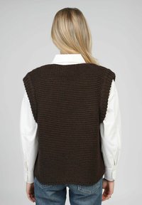 Veste marron tricotée avec une surface texturée, design à épaules larges et coupe décontractée, portée sur une chemise blanche à col.