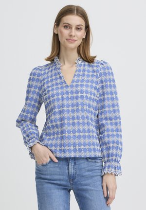 Joven mujer con blusa de estampado azul claro con cuello y puños fruncidos, y jeans azules, de pie con una mano en el bolsillo.