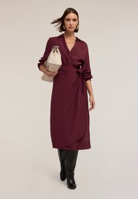 Eine Frau geht, trägt ein langes burgunderfarbenes Wickelkleid, schwarze kniehohe Stiefel, goldene Ohrringe und hält eine große beigere Handtasche.