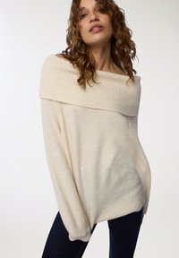 Maglione crema scollato con colletto a coste, texture morbida, maniche lunghe e vestibilità comoda, abbinato a jeans in denim scuro.