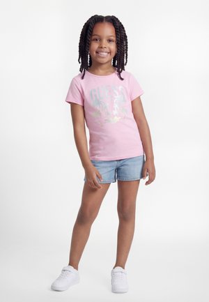 Jeune fille souriante aux cheveux tressés, portant un T-shirt rose à motif palmiers, un short en denim et des baskets blanches, debout devant un fond blanc.