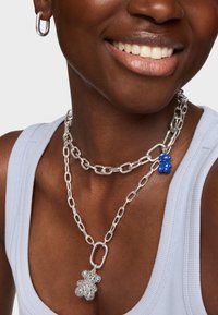 Collares de cadena de metal plateado superpuestos, con un encanto de oso azul. El modelo lleva una camiseta sin mangas de canalé azul claro y pendientes de aro.