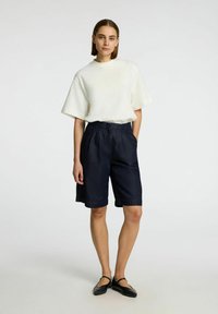 T-shirt blanche à manches courtes avec un logo discret, associée à un short bleu marine jusqu'aux genoux en tissu texturé et avec une taille élastique.