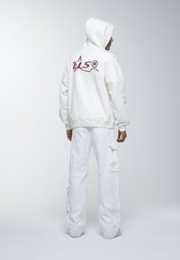Felpa bianca con un grande logo grafico rosso sulla schiena, abbinata a pantaloni cargo bianchi larghi con tasche laterali.