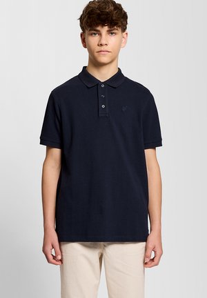Giovane uomo che indossa una polo blu navy a maniche corte e pantaloni beige, in piedi davanti a uno sfondo chiaro e uniforme.